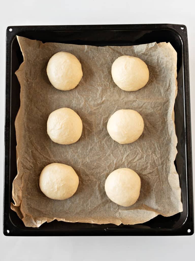 Parchment paper on a black baking tray with six burger buns before rising.