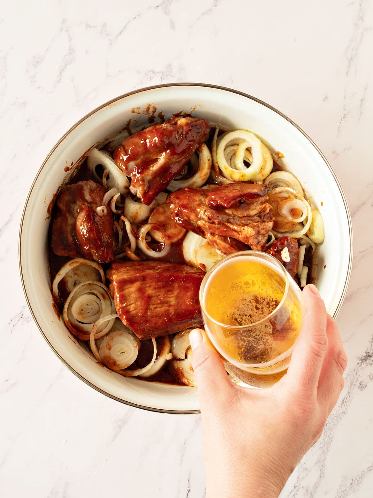 Adding beer to pork and onion pieces in a white saucepan. Marbled background.