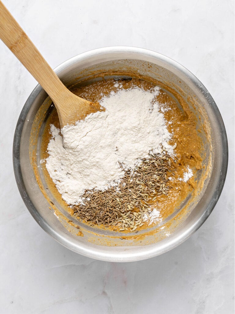 Flour and seeds added to rye bread mixture. Metal bowl, light gray surface.