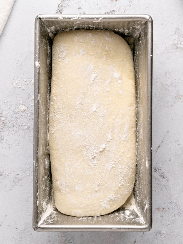 Loaf pan with white sandwich bread before rising. Light gray background.