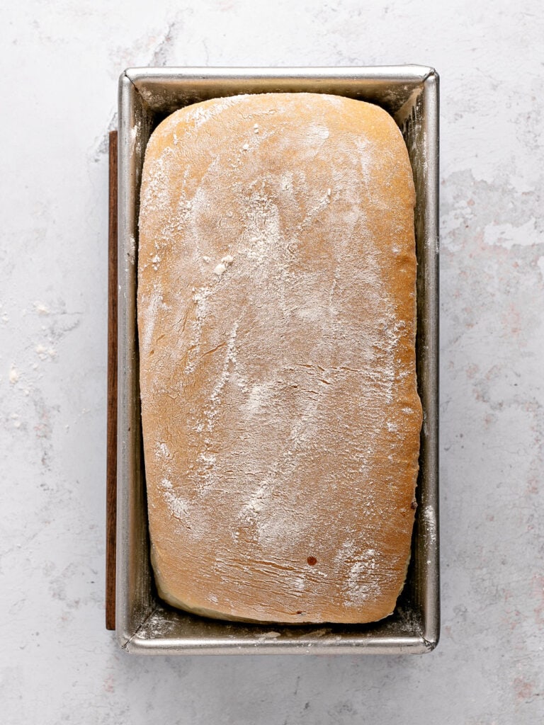 Baked white sandwich bread in a metal loaf pan. Light gray background.