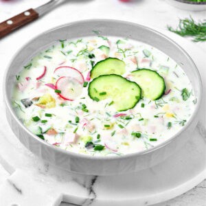 Light gray shallow bowl with cold cucumber, ham, kefir, radish soup. White background.