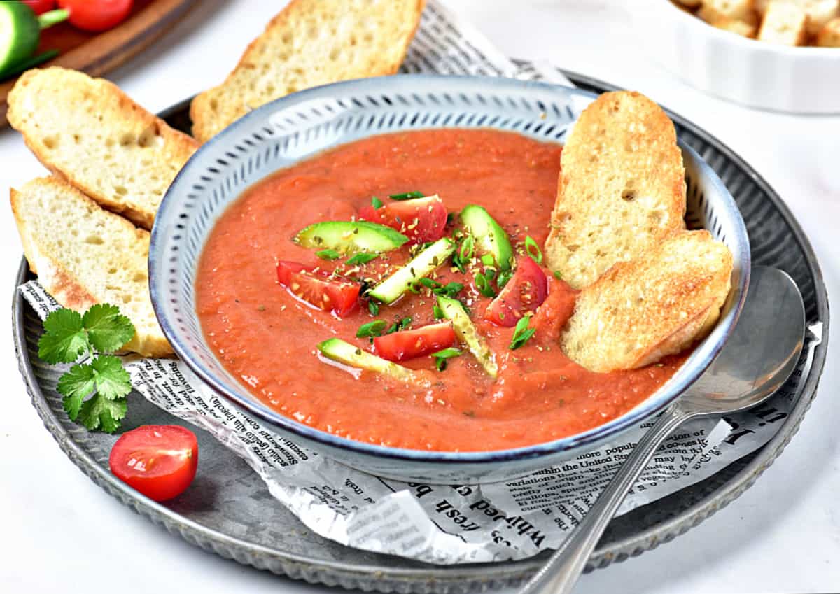 Gray bowl with gazpacho. Silver spoon and tray. Bread pieces.