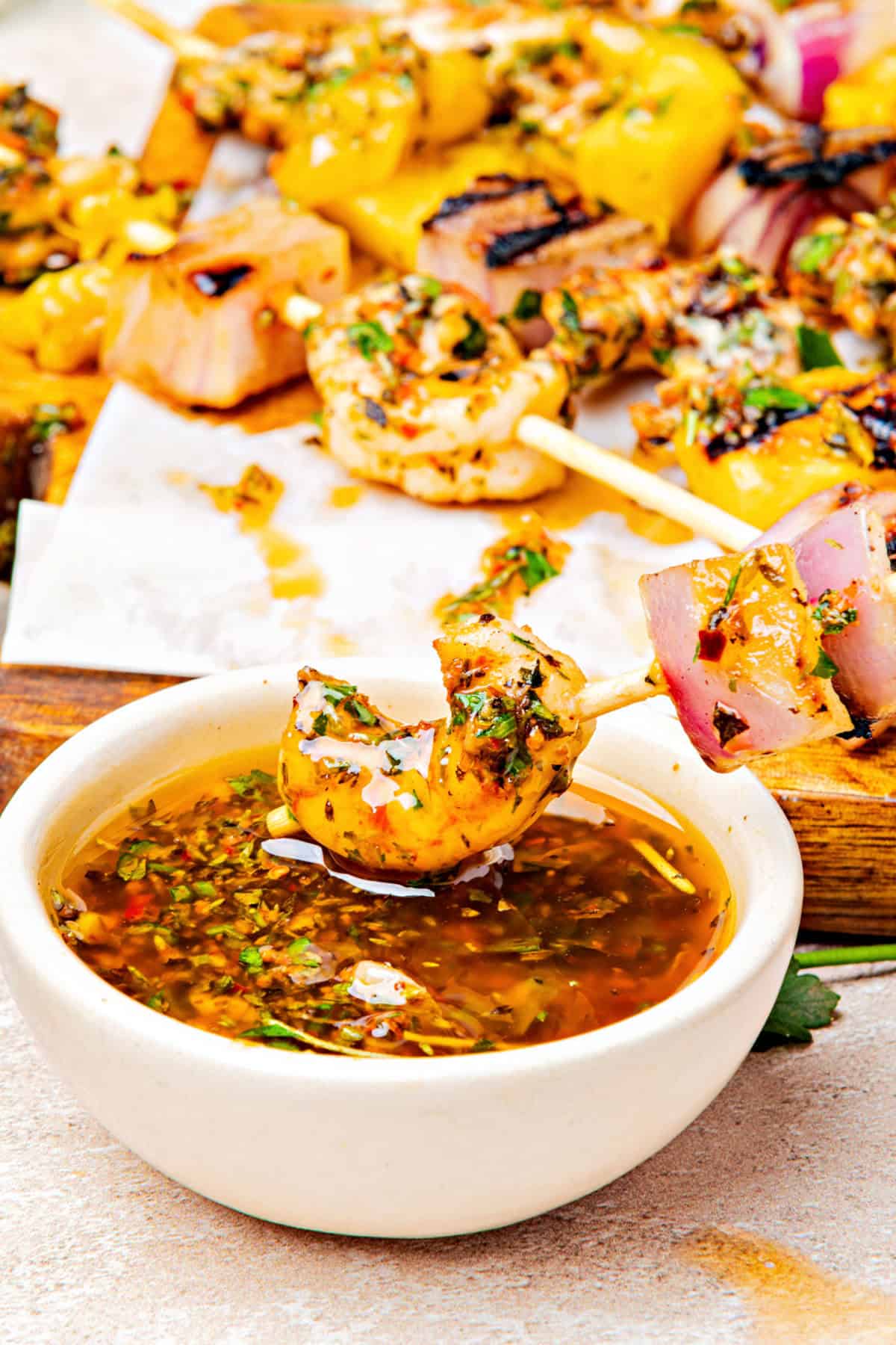 Dipping grilled shrimp skewer in a white chimichurri bowl. Skewers in the background.