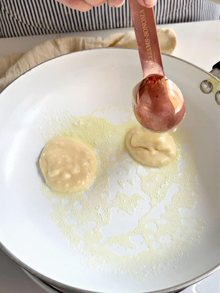 Pouring tablespoons of pancake batter on white skillet with melted butter.