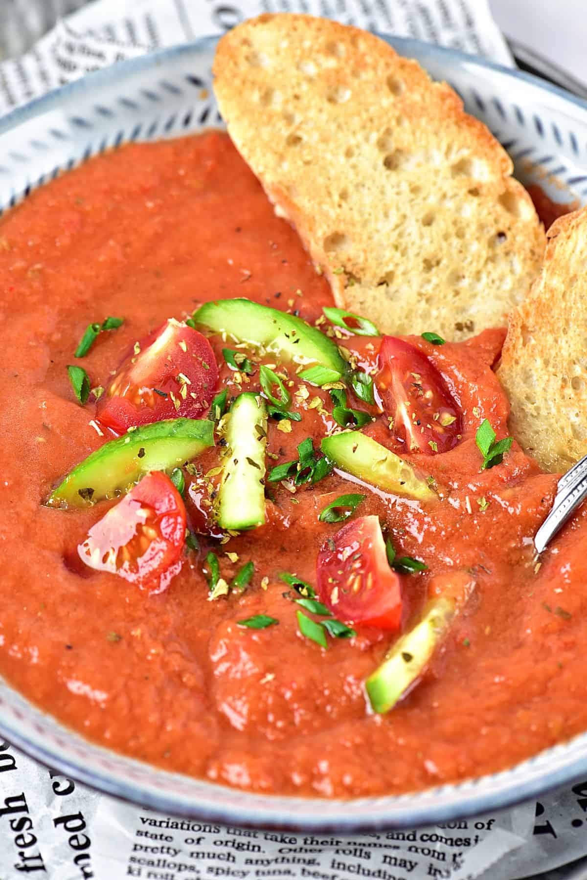 Gray bowl with gazpacho, bread pieces and garnishes.
