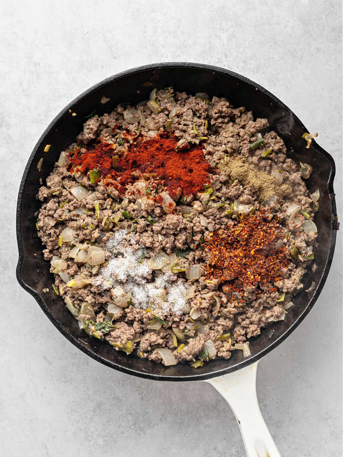 Seasonings and cooked ground meat in a black skillet. Light gray surface.