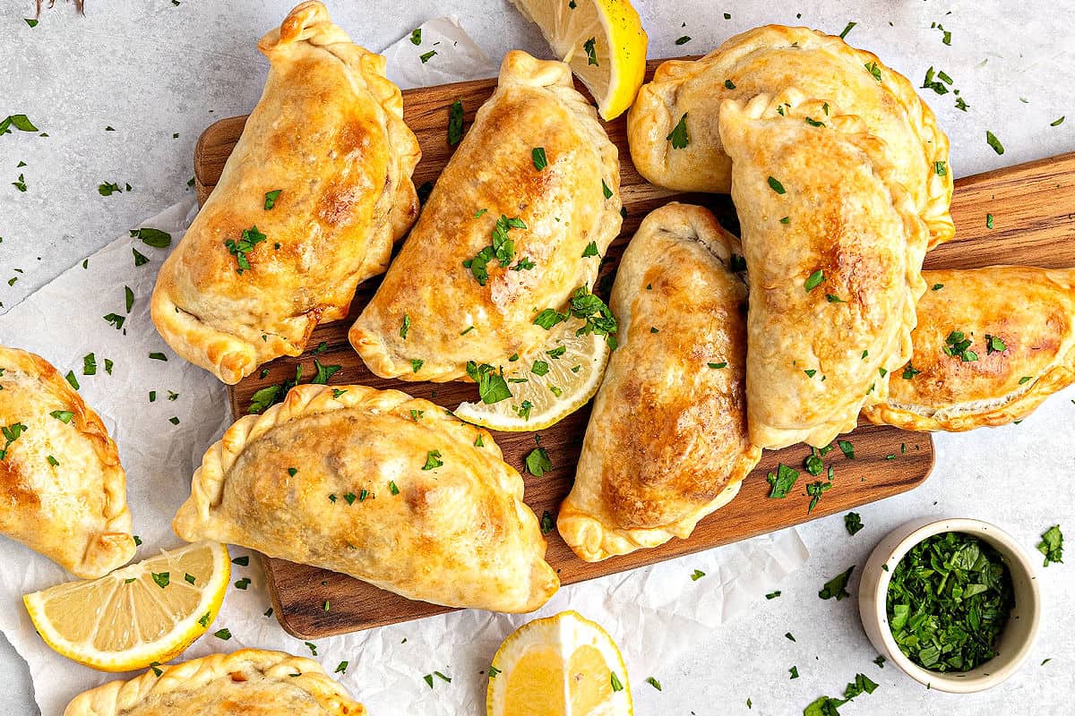 Wooden board with several Argentine beef empanadas. Chopped parsley, lemon wedges.