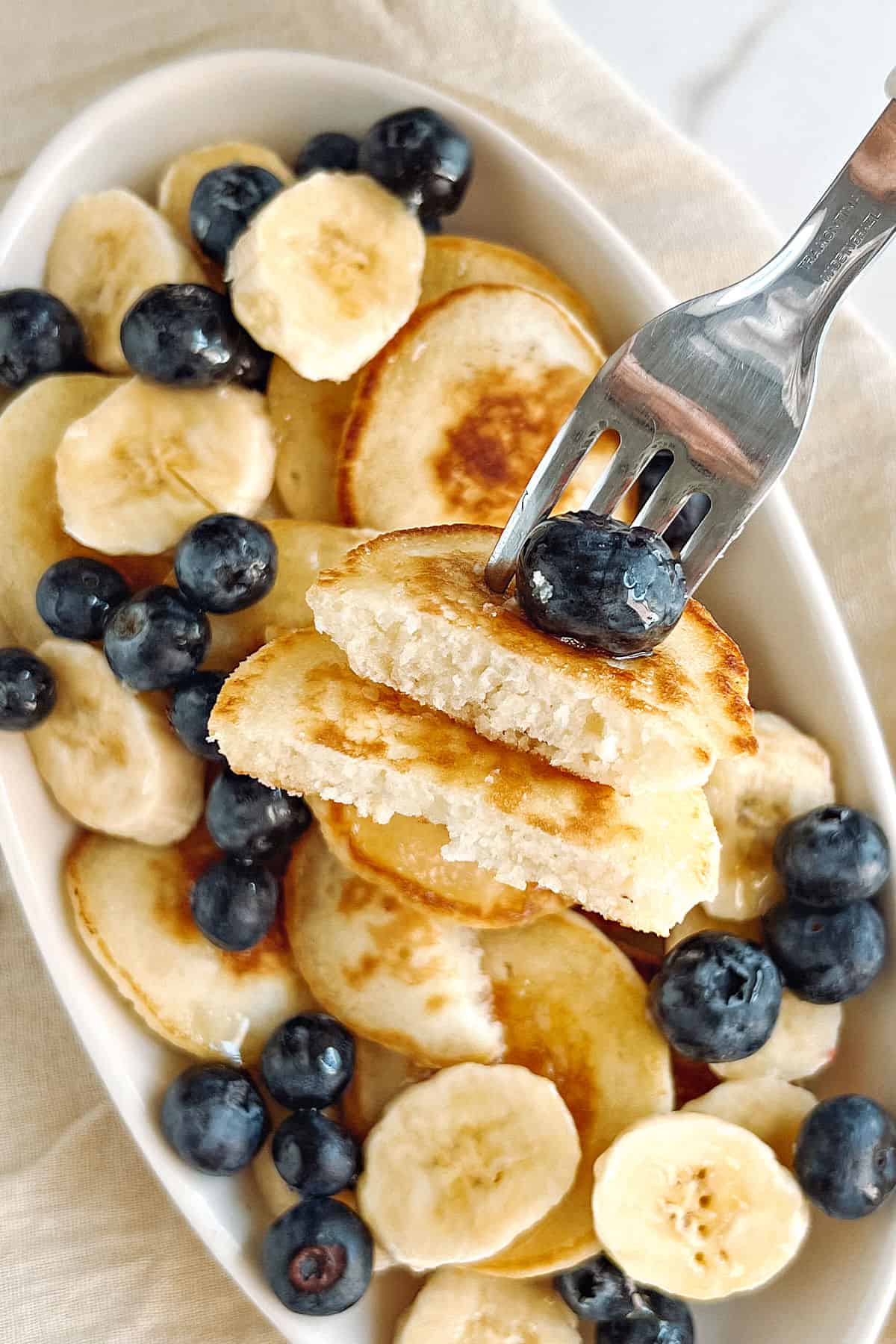 Fork halved silver dollar pancakes above dish with more pancakes and blueberries.