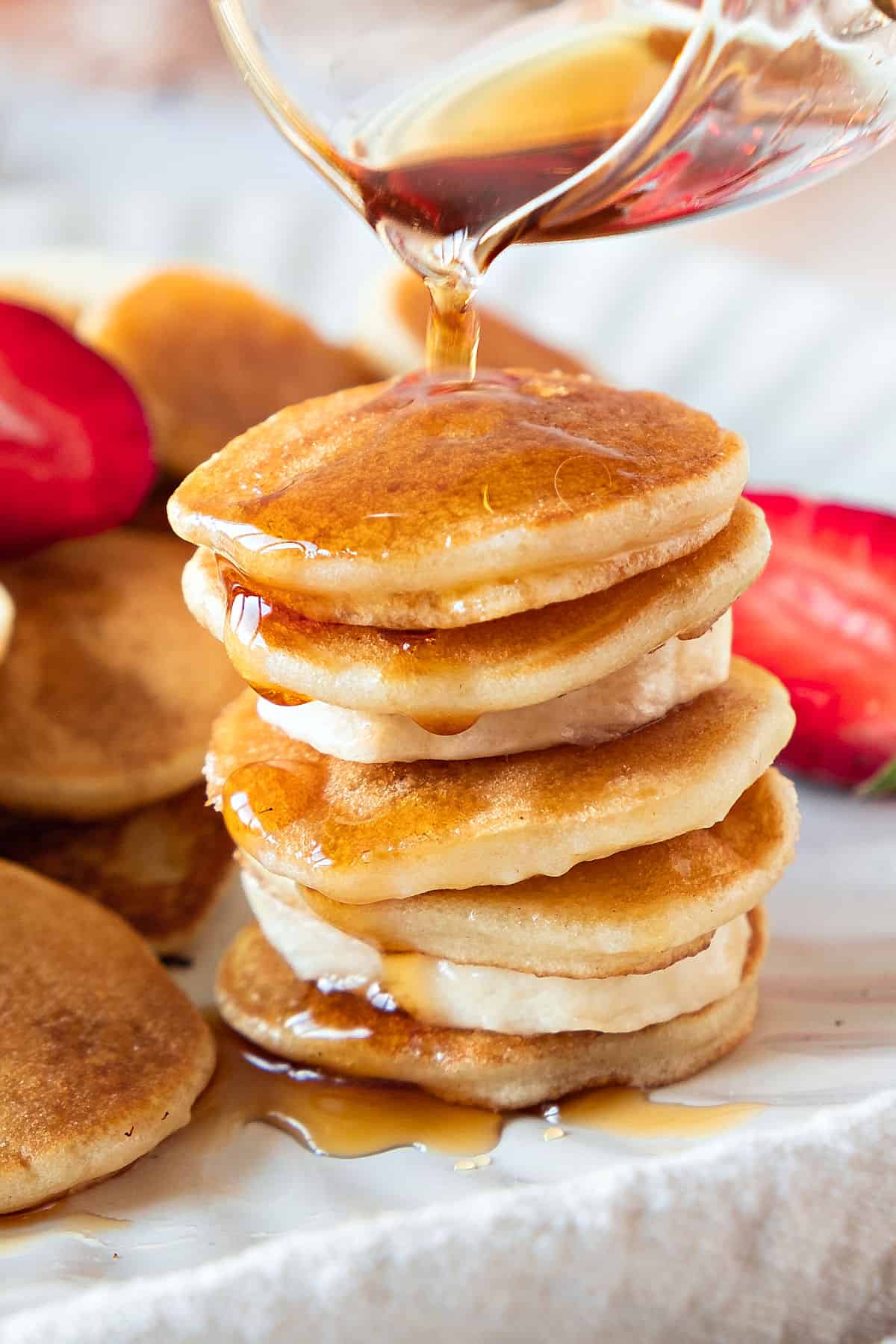 Pouring syrup on stack of mini pancakes and banana slices. White plate.