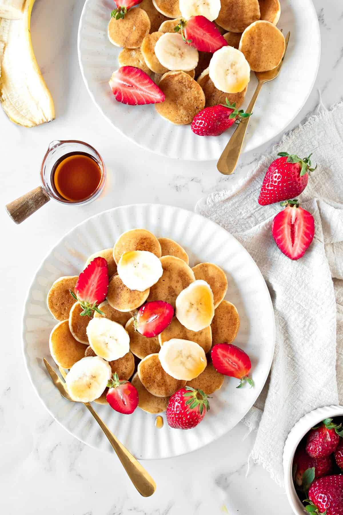 Top view of white plates of mini pancakes, banana and strawberries. White background.