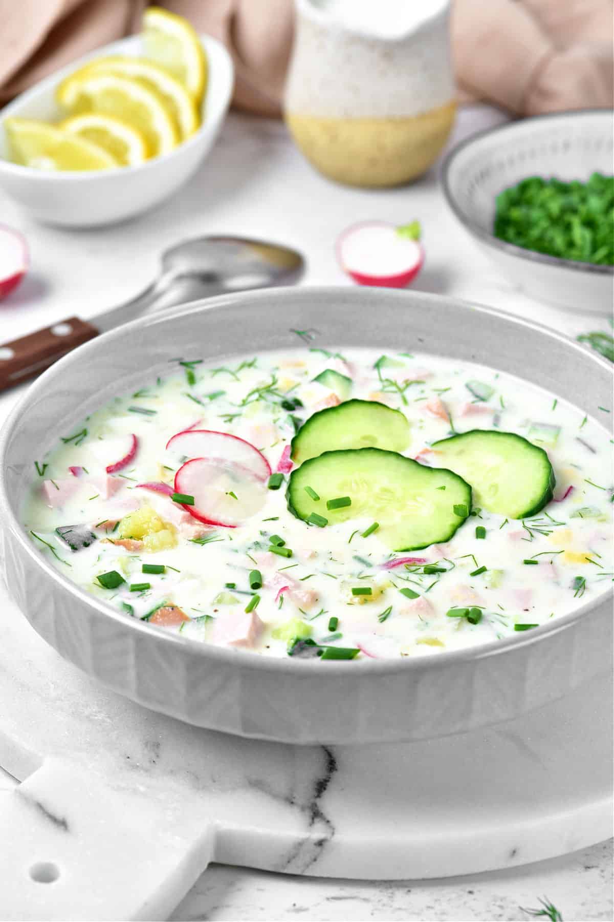Serving of cold cucumber, radish, ham, dill soup in a light gray bowl. White background.