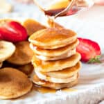 Stack of silver dollar pancakes with syrup, more pancakes and strawberry pieces.