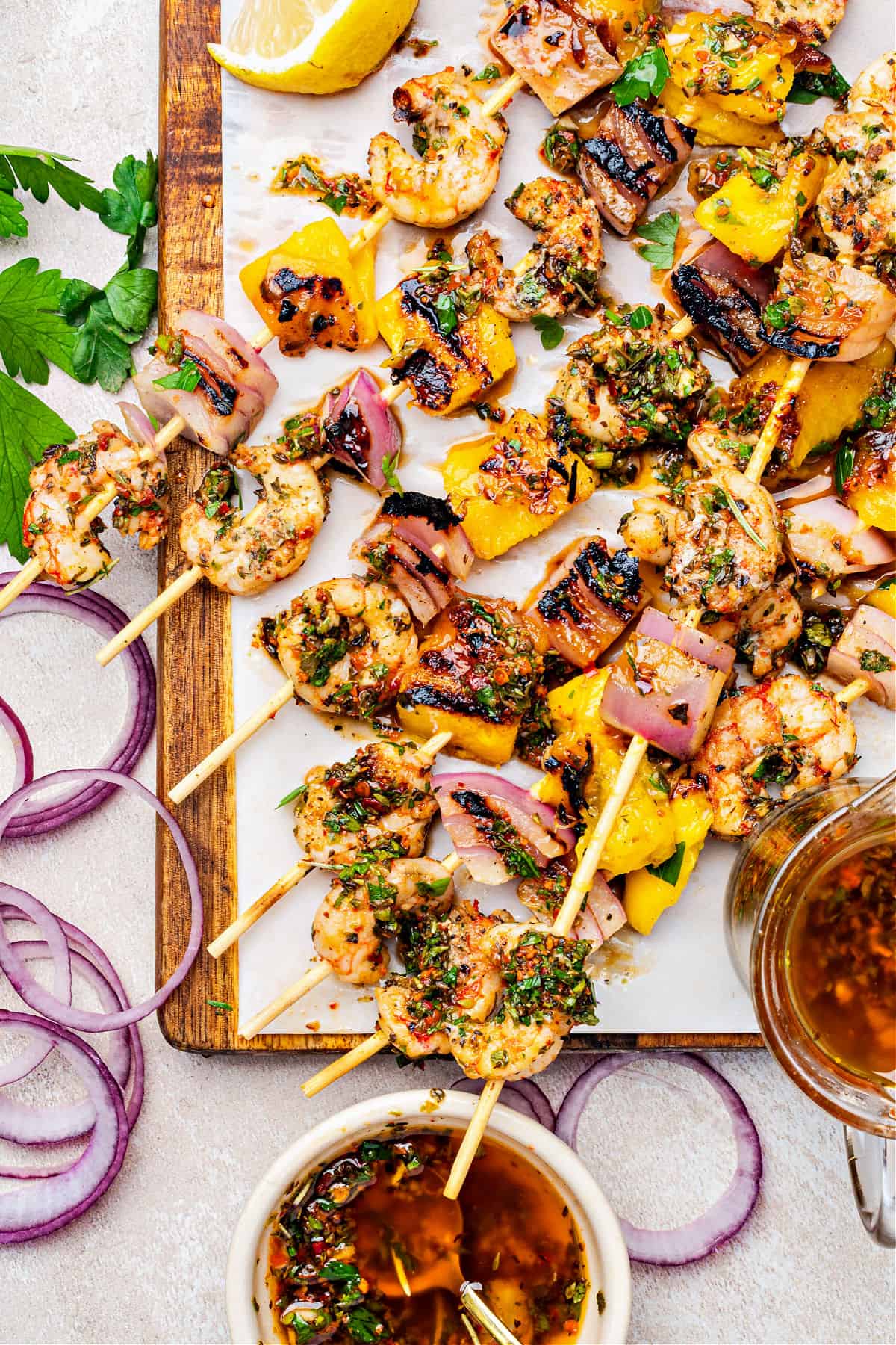 Top view of shrimp chimichurri skewers on a white paper and wooden board. Bowls of sauce.