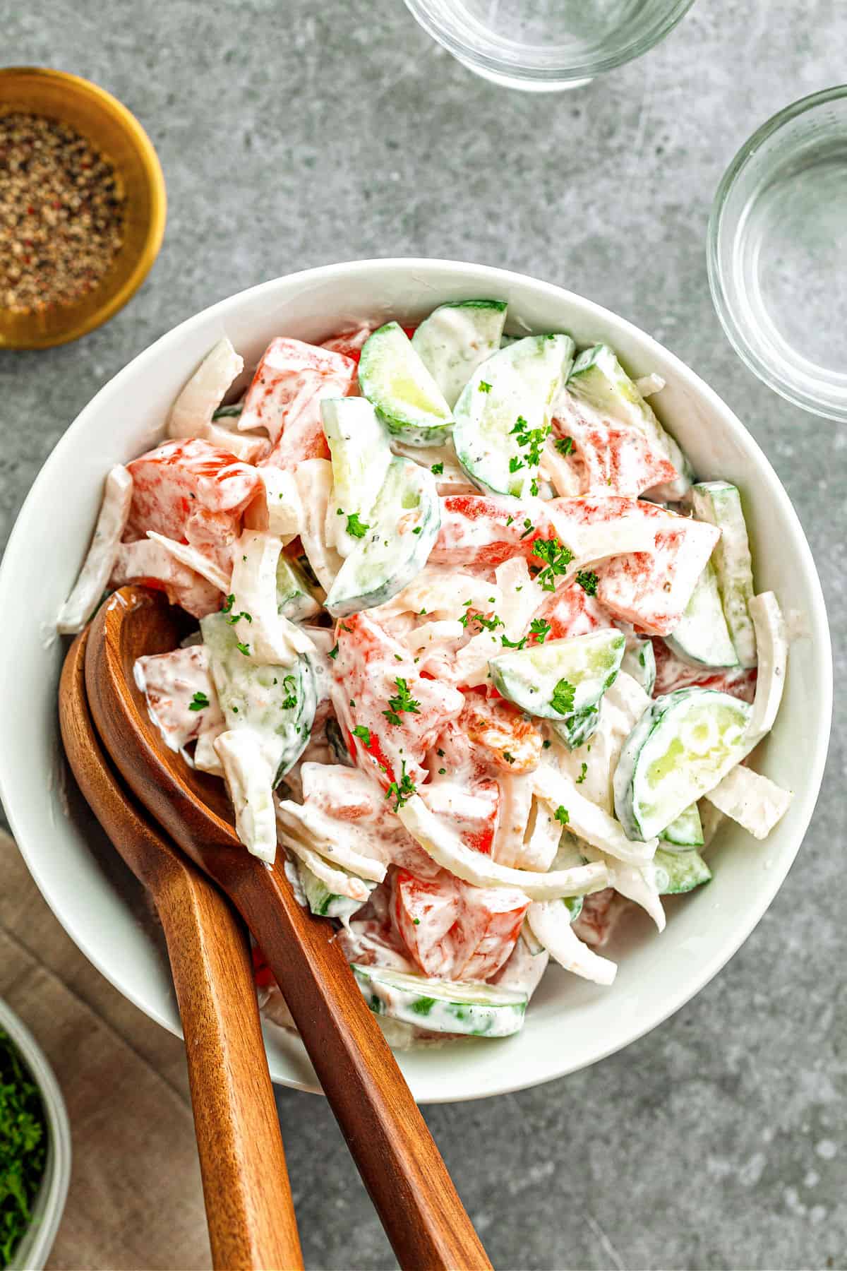 Creamy cucumber tomato salad in a white bowl. Wooden utensils. Gray background.
