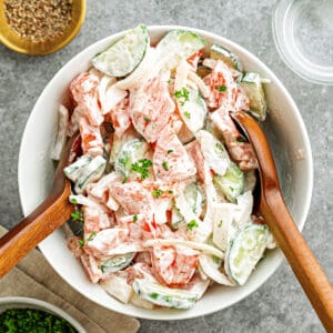 White bowl of creamy cucumber tomato salad. Wooden utensils. Gray background.