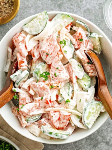 White bowl of creamy cucumber tomato salad. Wooden utensils. Gray background.