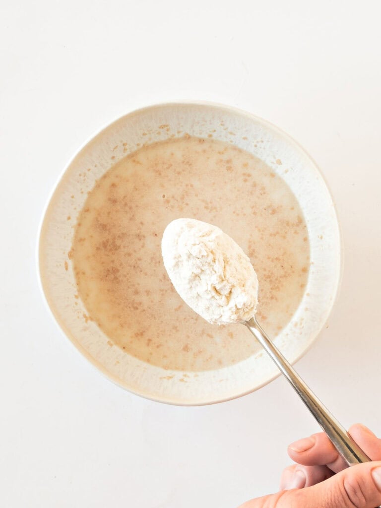 Adding flour to a white bowl with yeast and water.