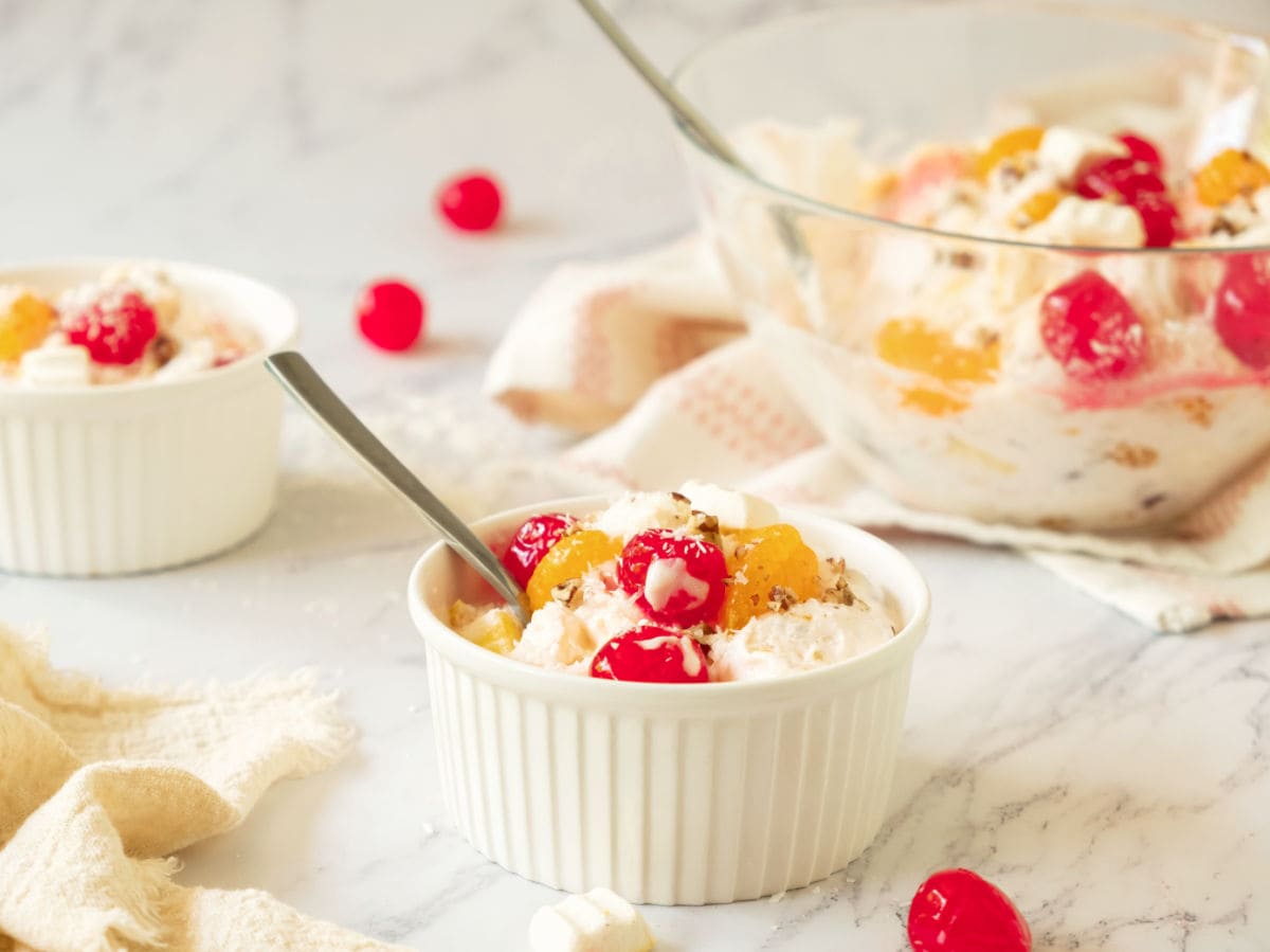 White and glass bowls with ambrosia salad and silver spoons. White marble background.