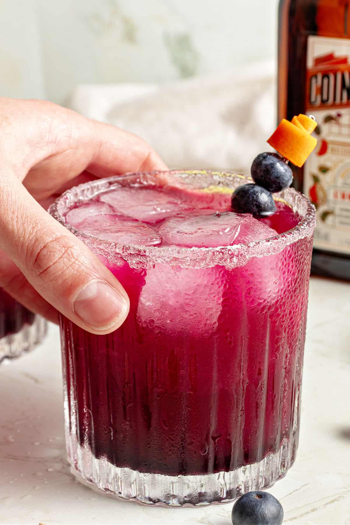 Hand holding blueberry margarita glass. White background, partial bottle.