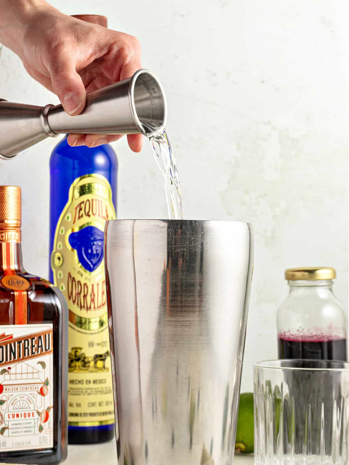Adding tequila to a metal shaker. Bottles in the light gray background.