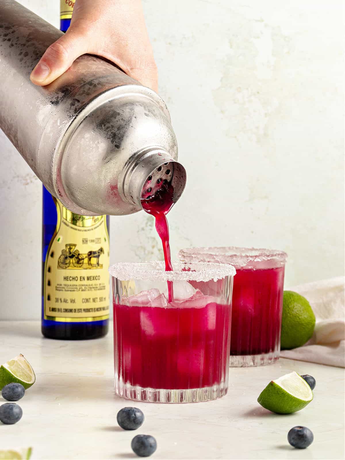 Pouring blueberry margarita from a shaker to a whisky glass. Light gray background.