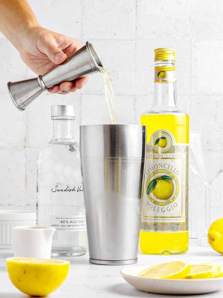 Pouring limoncello from a metal jigger to a shaker. Bottle, lemons slices, light gray background.