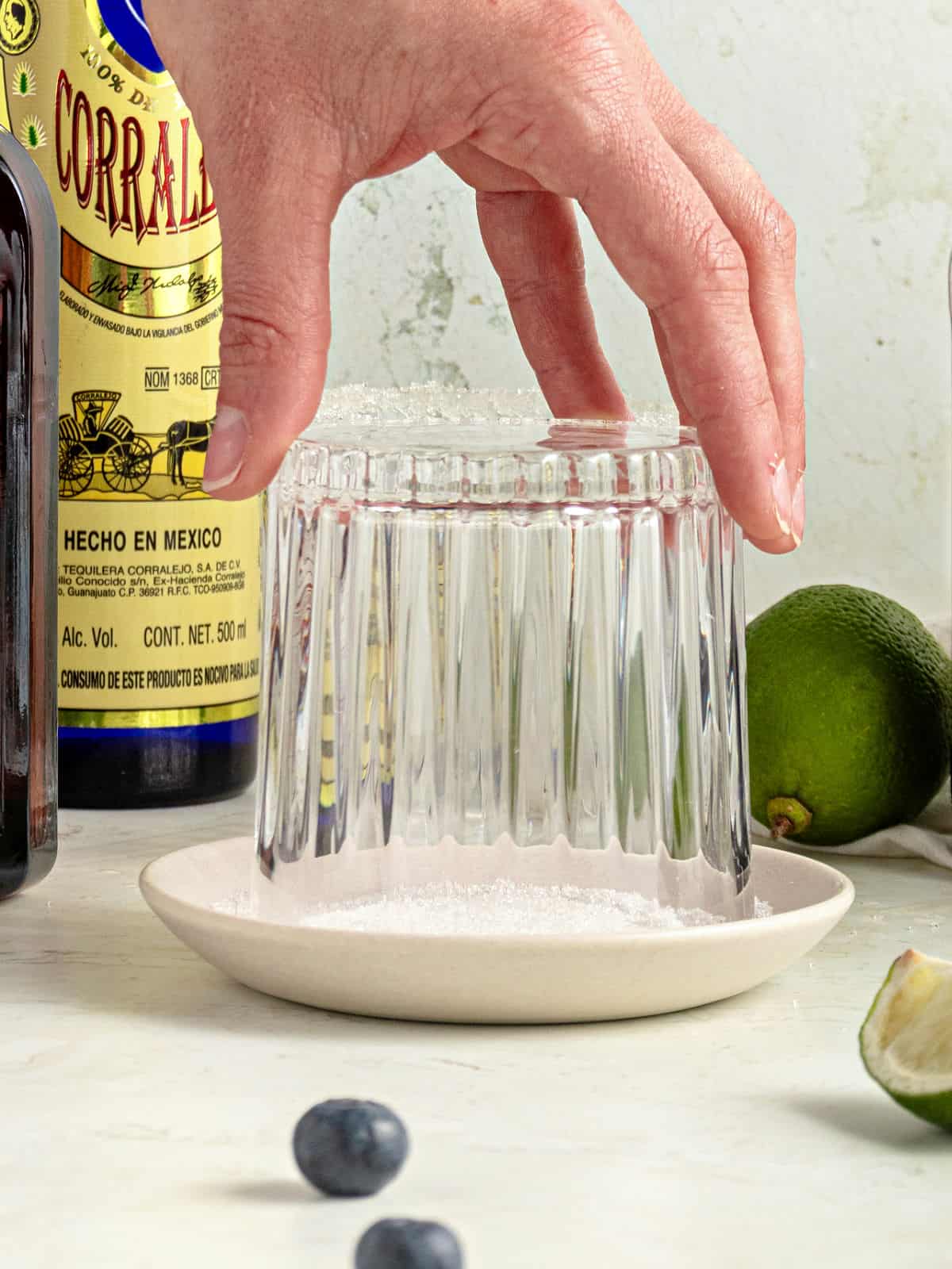 Placing a glass upside-down on a white plate with sugar. Light gray background. Bottle and limes.