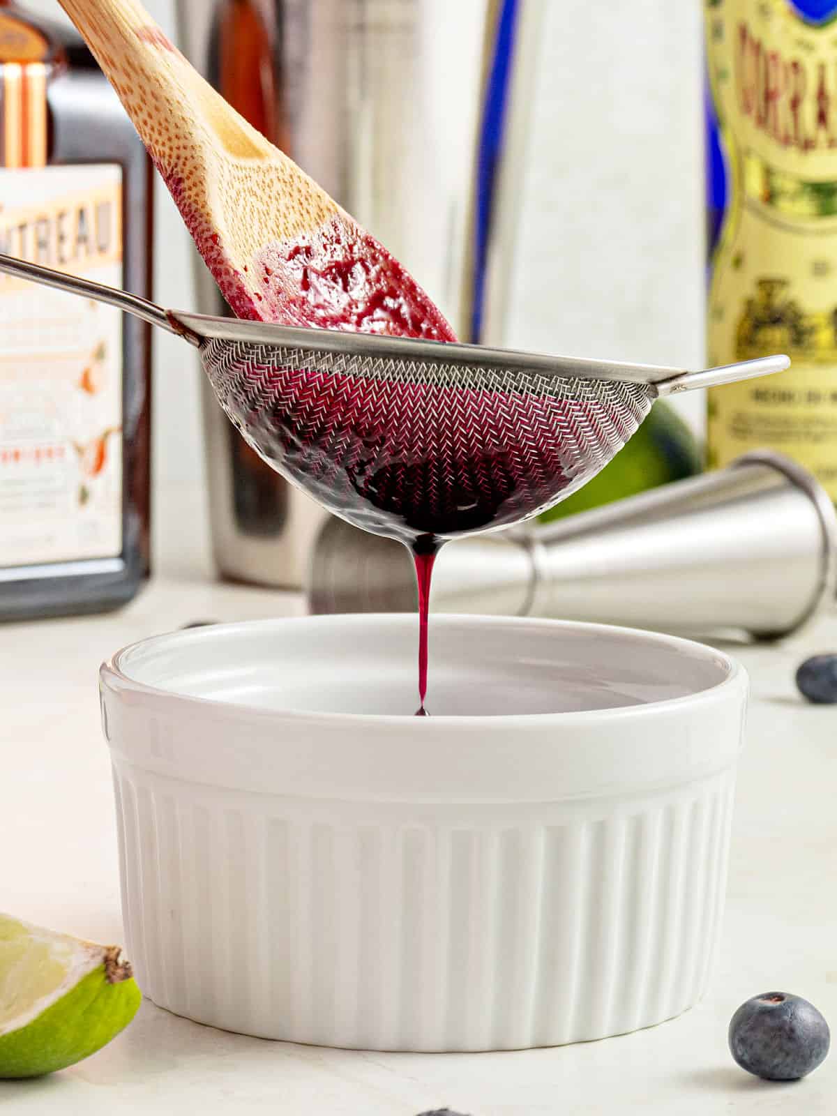Straining blueberry syrup over a white bowl. Bottles and shaker in the background.