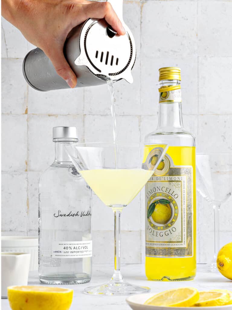 Pouring limoncello martini from a shaker to a martini stem glass. Light gray background.