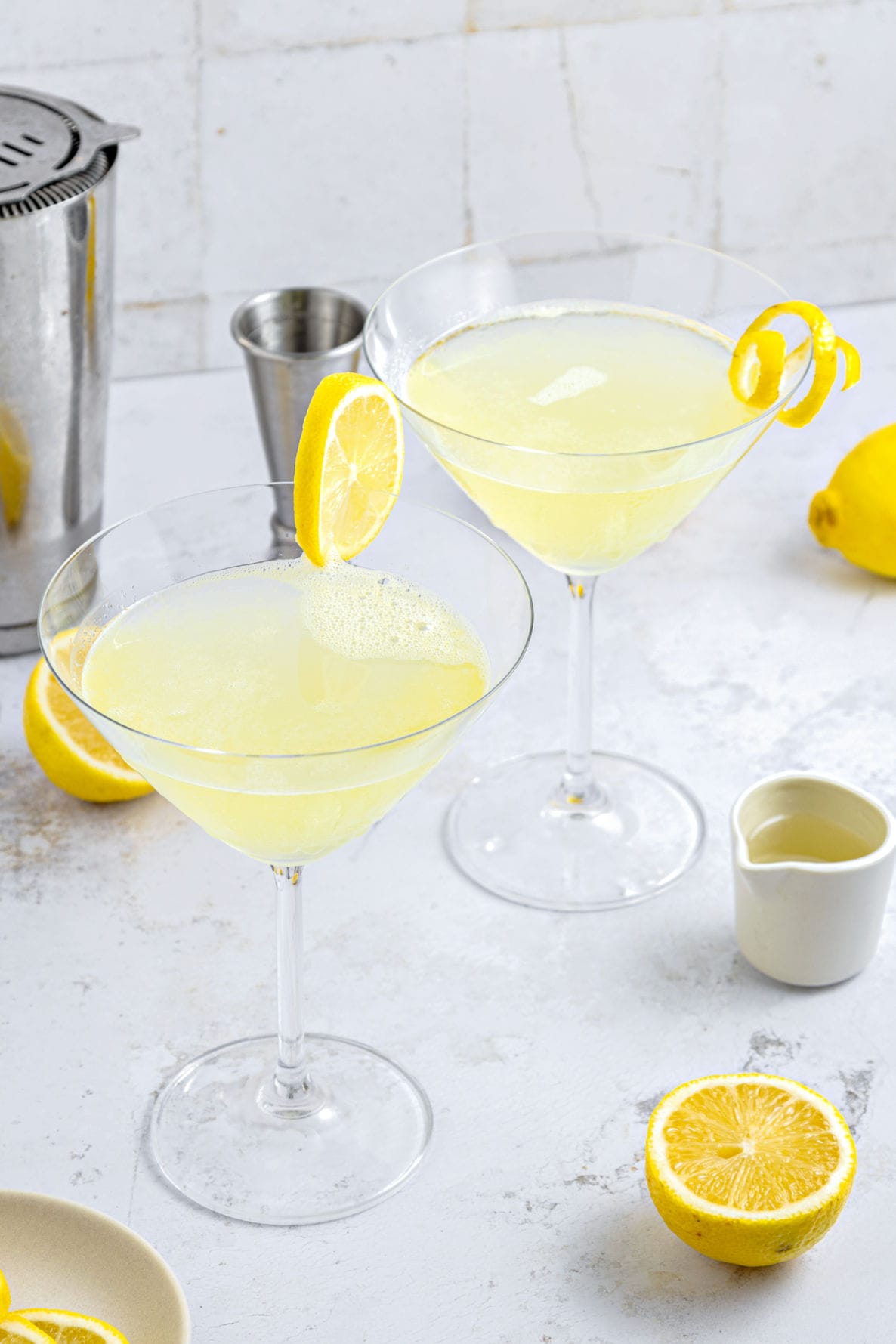 Light gray background with two lemon martinis. Shaker, jigger, lemon slices.