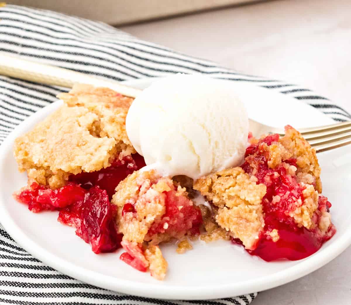 Serving of strawberry pineapple dump cake with ice cream on a striped cloth.