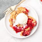 White plate with strawberry pineapple dump cake serving. Ice cream scoop, striped cloth, white background.