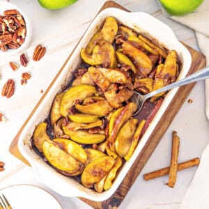 Cinnamon baked apple pieces in sryup in a white rectangular dish. White background, pecans, cinnamon sticks.