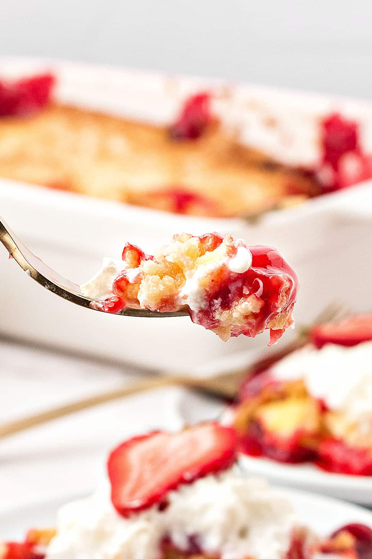Fork with bite of cream cheese strawberry dump cake. White dish and plates with more cake below.