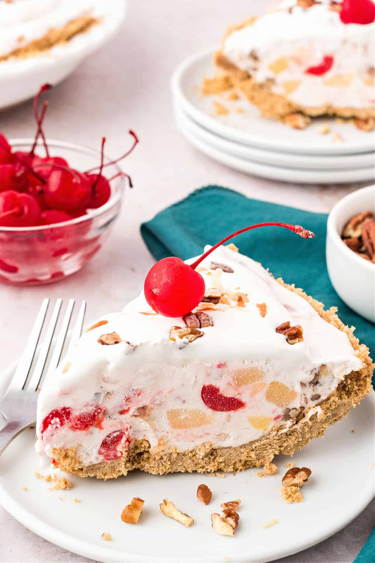 Slices of millionaire cream pie on white plates. White background, bowl of cherries, teal kitchen towel.