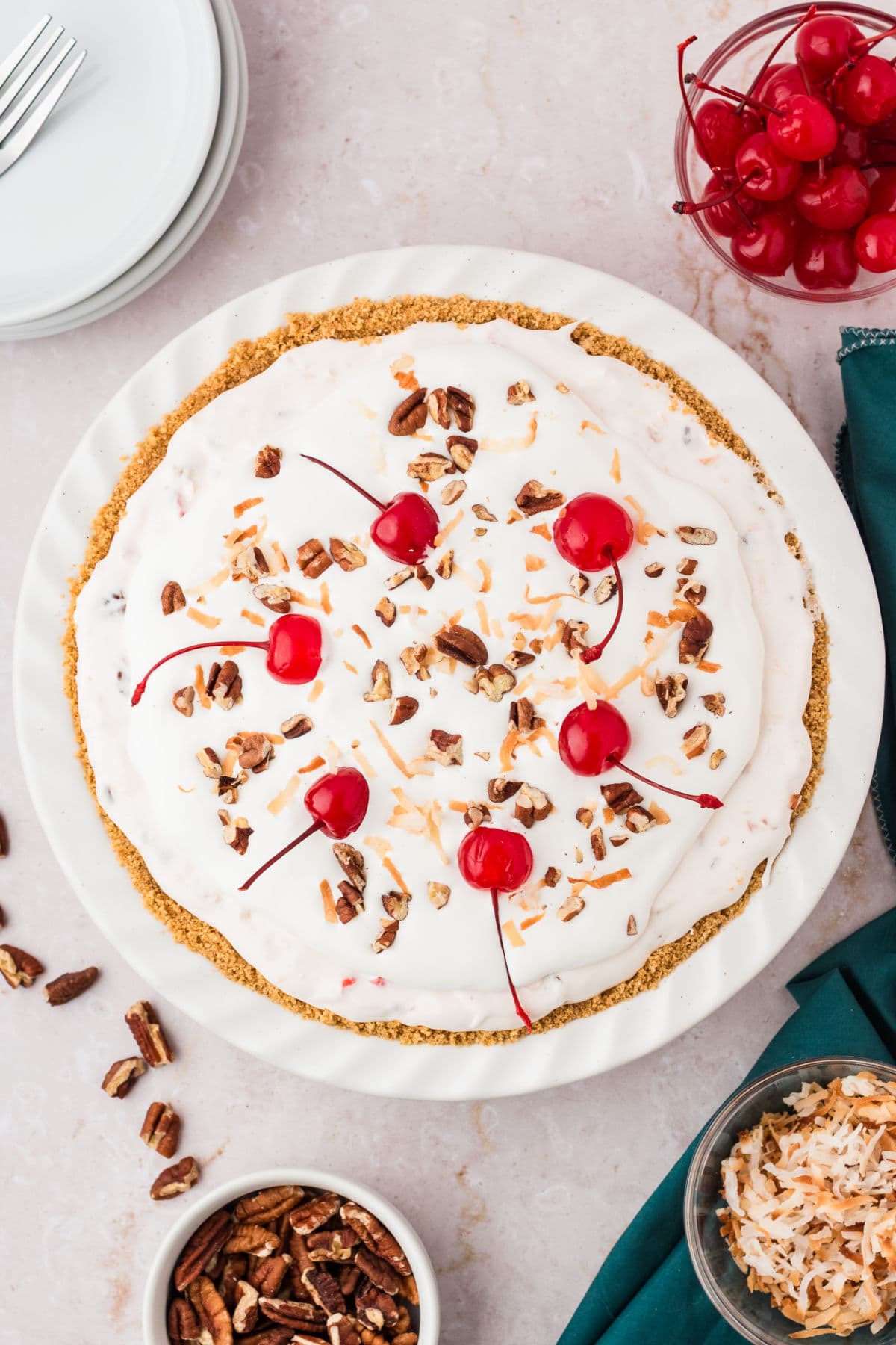 Whole cherry pecan topped cream millionaire pie on a white plate. Whitish background. Cherries, pecans, teal cloth.