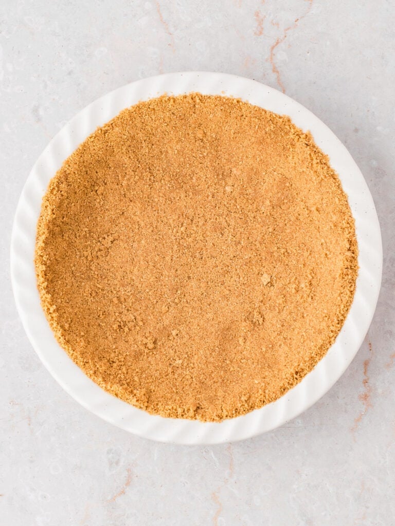 Graham cracker crust in a white pie plate. Light gray background. Top view.