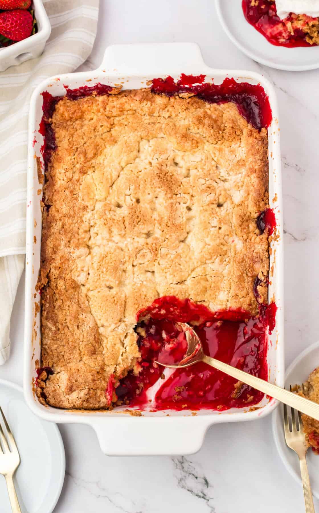 White dish with strawberry dump cake, serving missing. Silver spoon, white marbled surface, top view.