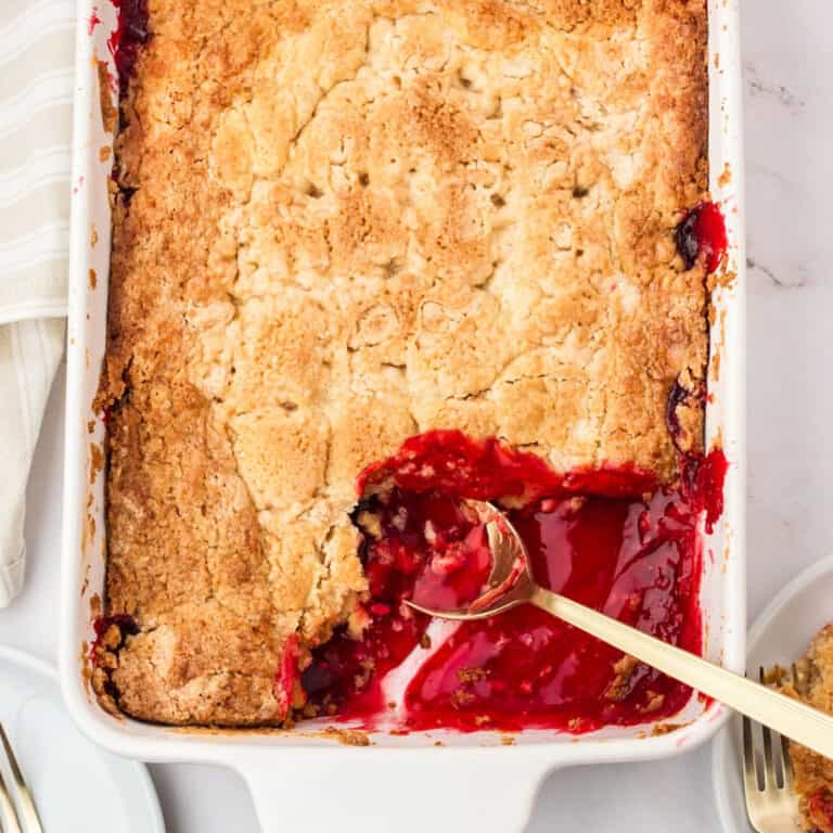 Partial view of white dish with strawberry dump cake, serving missing. Silver spoon.