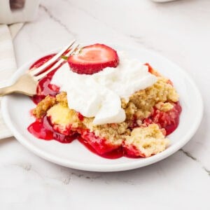 White plate with serving of cream-topped strawberry dump cake. White marbled surface.