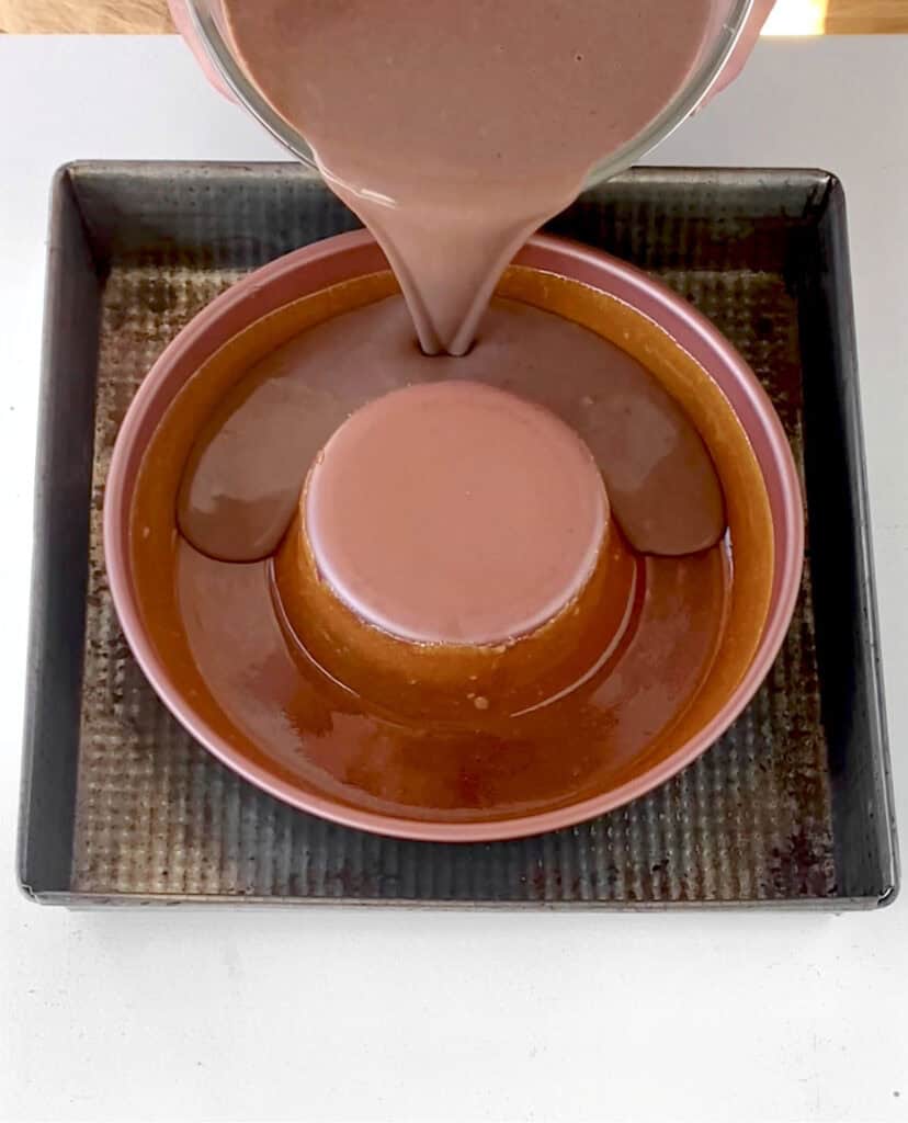 Pouring chocolate flan mixture into a caramelized copper tube pan inside a metal pan.