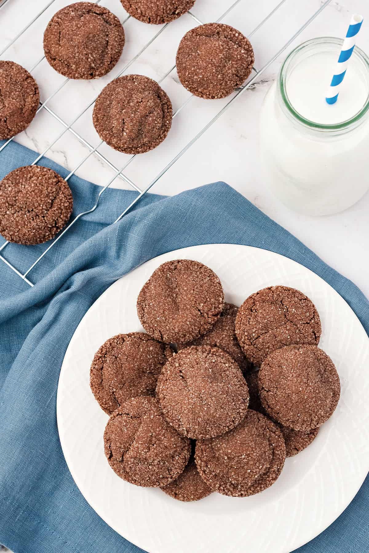 Top view of white plate with pile of chocolate cookies. More on a wire rack. Blue towel. 
