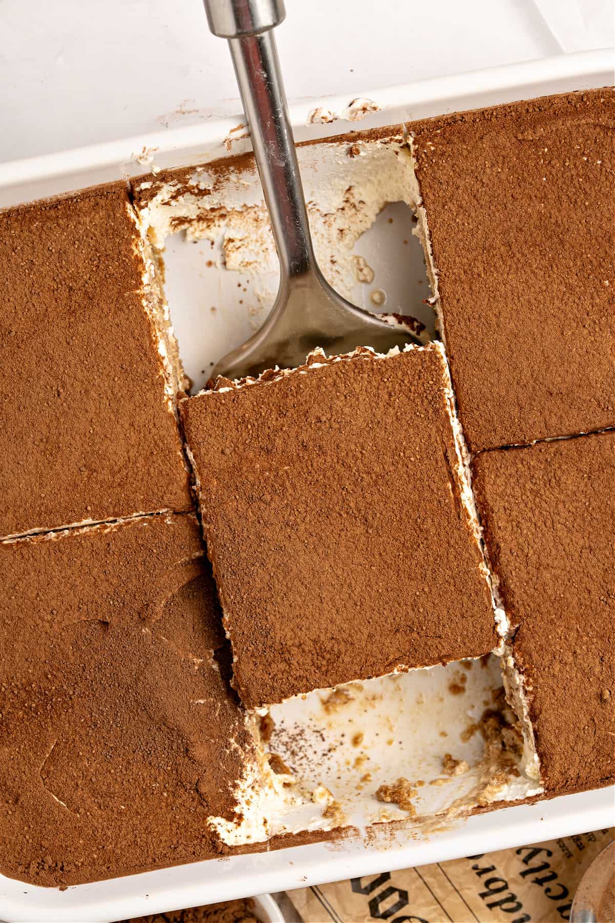 Metal spatula with tiramisu serving in a white dish with the remaining dessert.