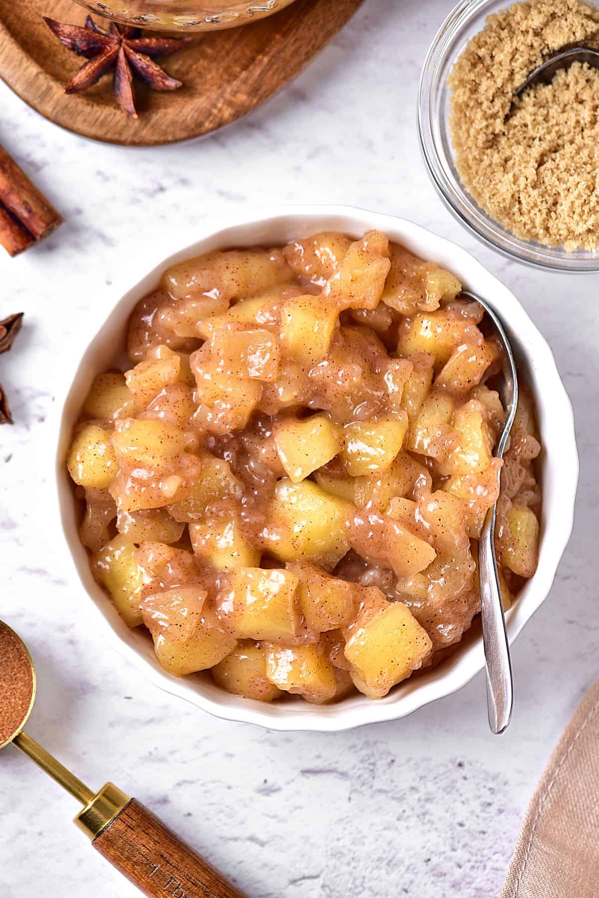 Round white dish with apple topping and filling. White background. Cinnamon sticks, brown sugar.
