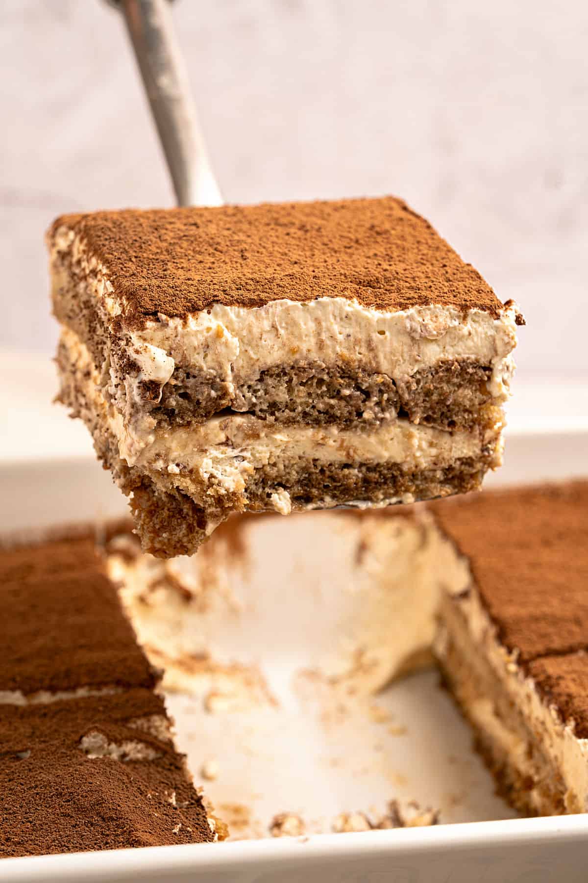Lifting a serving of tiramisu from a white baking dish. Light gray background.