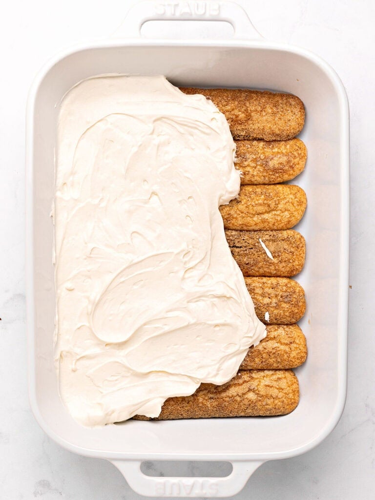 Spreading mascarpone filling over soaked savoiardi biscuits. White rectangular dish.