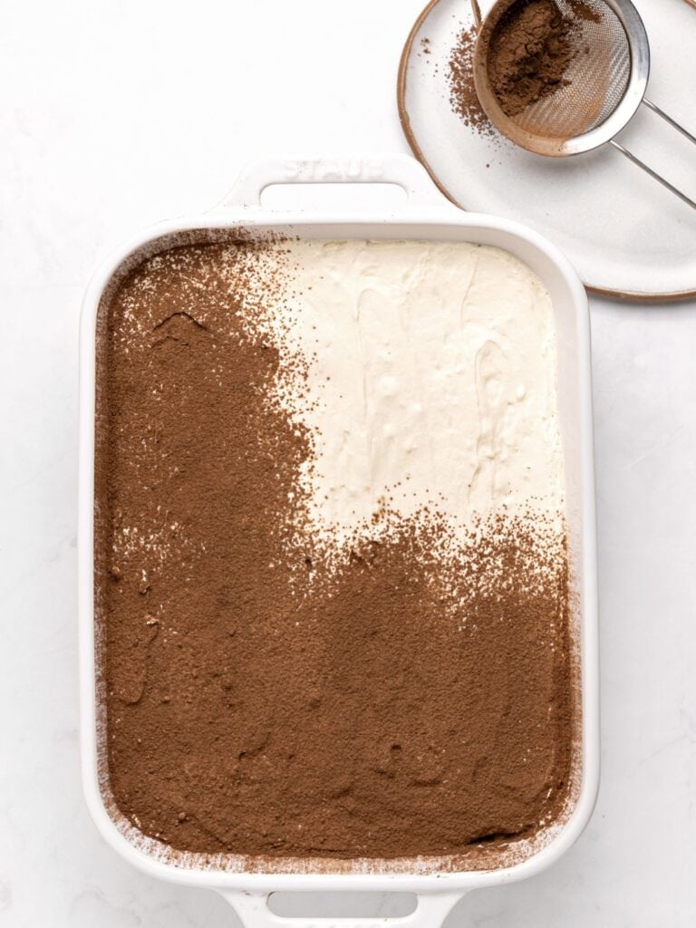 Dusting cocoa powder on mascarpone layer in a white dish. Small sifter with cocoa.