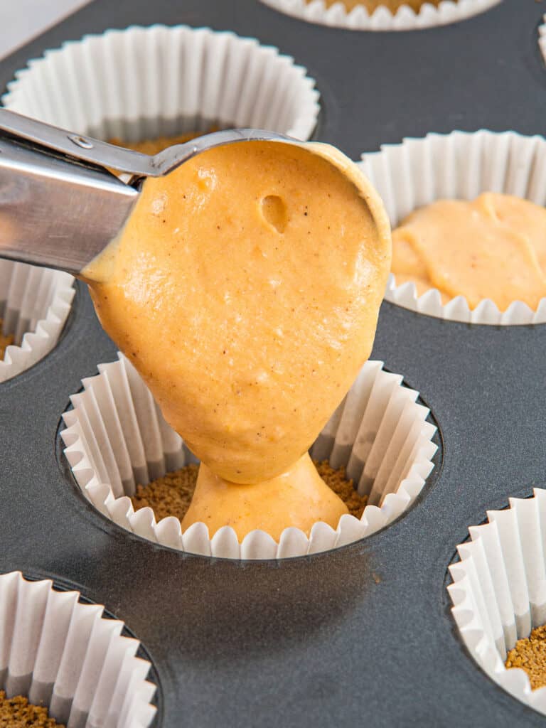 Scooping pumpkin cheesecake filling into mini paper liners. Metal pan.