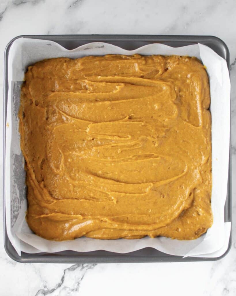 Parchment-lined square metal pan with pumpkin blondie batter. Marbled surface. 